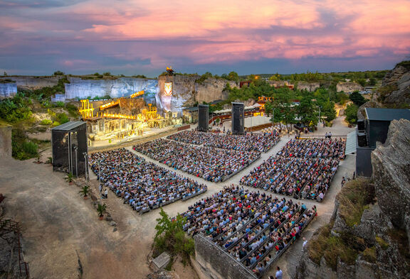 2022 Steinbruch St Margarethen | Andreas Tischler Menschen sitzen auf Tribünen und genießen ein lebhaftes Open-Air-Konzert in der Natur.
